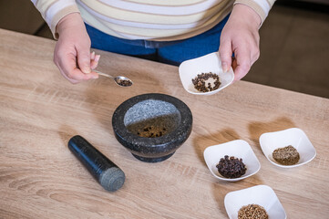 A woman's hand pours spices into a marble mortar. In the bowls are the spices: black pepper, zira and mustard seeds.