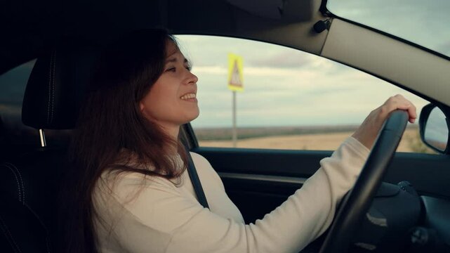 Happy woman drives car looking in rear view mirrors driving along rural road. Positive woman drives modern car across countryside on vacation. Woman driver controls car having trip across farmland