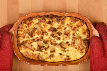 A freshly baked strata (layered casserole dish made from a mixture which mainly consists of bread, eggs and cheese) is being held with oven mitts on a wooden surface