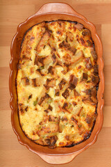 A  baked strata (layered casserole dish made from a mixture which mainly consists of bread, eggs and cheese) in a rustic dish sits on a wooden table, ready to be served