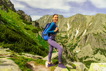 Naklejka premium Excited woman tourist woman with backpack walking along hiking trail against the backdrop of beautiful mountain scenery. Travel, vacation, active lifestyle