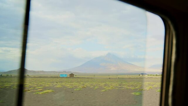 View of Ol Doinyo Lengai volcano in Tanaznia during drive from safari car window. Driving in Africa for safari tour looking out from window during ride or game drive. Tall mountain in background