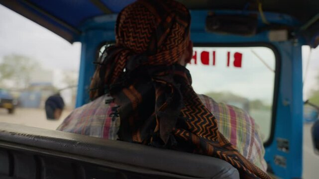 Man driver rides blue auto-rickshaw tuk tuk in African village. Wears patterned scarf and checked shirt. Navigates through rural countryside. Everyday transport in small village or town in Tanzania