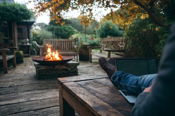 Person Works on Laptop Outdoors With Fire Pit, Relaxing on Wood Deck Amidst Lush Green Garden
