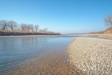 Calm river scene under a clear blue sky, with a rocky shore and leafless trees in the background.