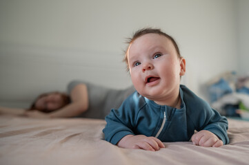 A three-month-old boy lies on his stomach on the bed and his mother sleeps behind him. Postpartum...