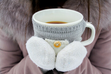 Hands in cozy coat hold a white mug with a grey knitted warmer. The image evokes feelings of warmth, comfort, and relaxation on a cold day.