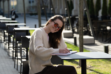 Fototapeta premium A woman sits at an outdoor table, smiling while writing in a notebook. The cafe is surrounded by trees and modern furniture in a sunny, tranquil setting
