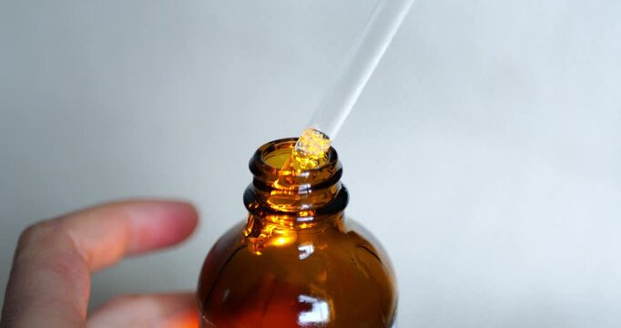 Hand holding a glass pipette with clear liquid above an amber dropper bottle. Macro shot on a plain background, ideal for concepts of essential oils, medicine, skincare, wellness, or science.