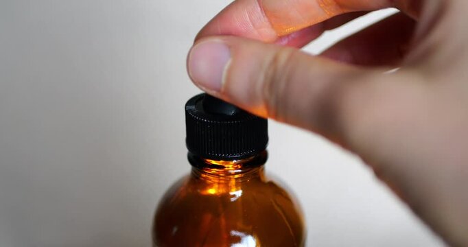 Hand opening a black dropper cap on an amber glass bottle with a plain background. Macro shot suitable for themes of essential oils, medicine, skincare, laboratory, wellness, and healthcare routines.