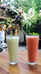 Two refreshing fruit smoothies in tall glasses with metal straws on a wooden table, served in a cozy outdoor café with a green natural background