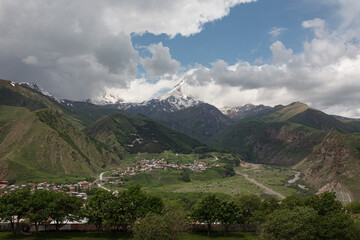 Fototapeta premium Scenic view of a mountain valley with a small village nestled among green hills, surrounded by towering peaks under dramatic clouds, creating a serene yet dynamic landscape.