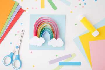 Handmade paper rainbow with white clouds made of colored paper strips on blue square background....