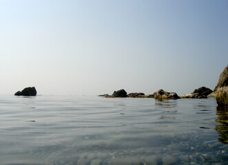 Hot, windless weather at sea. Sky and water almost merge. A small group of low rocks separates the sea and sky. The bottom of rounded stones shines through the calm water