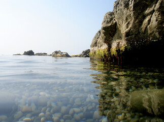 Hot, windless weather at sea. Sky and water almost merge. A small group of low rocks separates the sea and sky. The bottom of rounded stones shines through the calm water