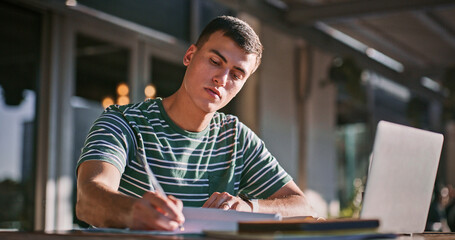 Man, university student and outdoor with laptop or writing on notebook for assignment on campus. Male person, college learner and textbook for education with research or study for exam and assessment © Mitchell/peopleimages.com