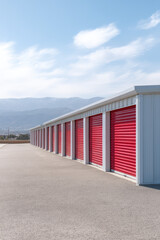 Obraz premium Row of storage units with vibrant red doors, situated in an outdoor facility. The sky is clear with a few clouds and mountains 