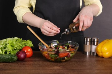 Woman pouring oil onto tasty salad at wooden table against dark background, closeup