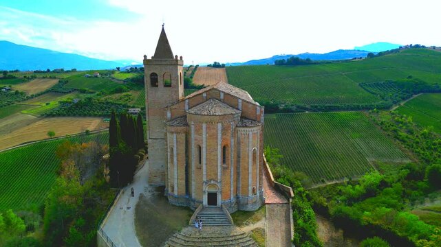 OFFIDA, ITALY - AUGUST 4, 2024: Aerial truck right shot of Chiesa di Santa Maria della Rocca. Visitors gather by the church, surrounded by sumptuous green hills and peaceful countryside views
