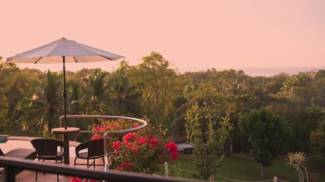 Outdoor terrace with round table and chairs under large parasol surrounded by tropical flowers and lush greenery during golden hour sunset with ocean view in distance