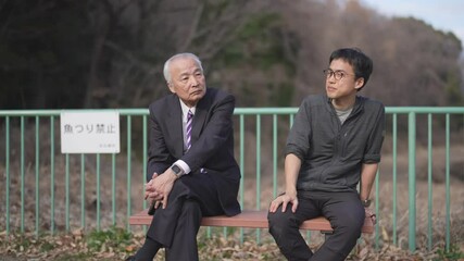 In a spring park, a man in his 70s in a suit and a man in his 30s sit on a bench talking. Their movements are captured in a slow-motion video surrounded by greenery and the peaceful spring air. - Powered by Adobe