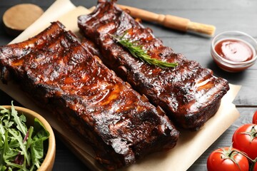 Tasty roasted pork ribs served with rosemary, tomatoes, sauce and salad on black wooden table, closeup