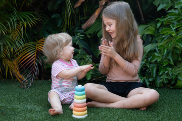 Happy sisters playing together on green lawn, baby clapping hands with older sister, joyful childhood moment in a tropical garden, natural light, colorful toy, family bonding, summer vibes