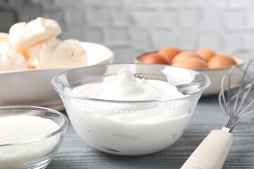Whipped cream in bowl, sugar, eggs and whisk on gray wooden table, closeup. Cooking meringue cookies