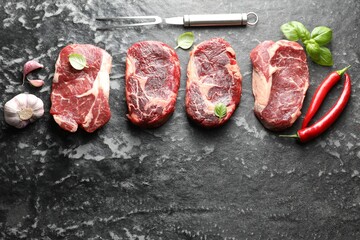 Pieces of raw beef meat, spices and carving fork on dark table, flat lay. Space for text