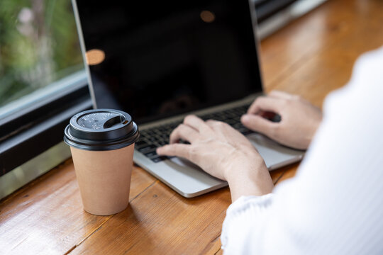 Male hands typing on laptop keyboard at coffee shop wooden table beside takeaway coffee cup. Modern freelance working lifestyle, digital technology concept, warm natural daylight.