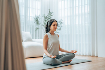 Young Asian woman meditating in lotus position at home, sitting on the floor in fitness clothing....