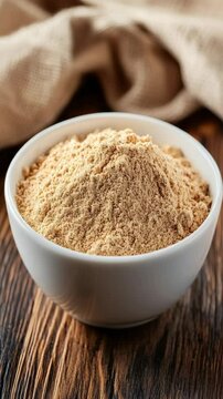 Nutritious maca powder in white ceramic bowl set against textured brown wooden surface with draped cloth backdrop.