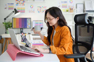 Young creative professional in casual workspace using laptop and sipping coffee, surrounded by colorful design charts.