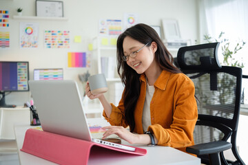 Young creative professional sitting at desk, holding a mug and looking at laptop in a bright, color-filled workspace.