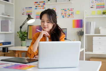 Focused woman using a laptop and stylus pen in a vibrant studio filled with color palettes, design...