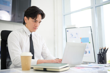 Obraz premium Focused young man in formal attire using a laptop at a modern office desk with coffee and documents.