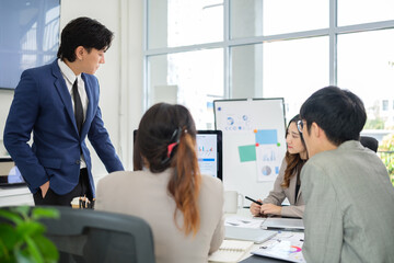 Group of business professionals gathered around a desk during a meeting, discussing charts and data on a computer and whiteboard.