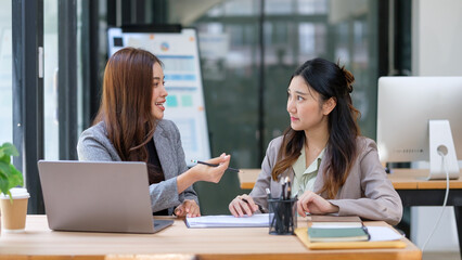 Businesswomen sitting at a desk in a modern office, engaged in a focused conversation with documents and a laptop