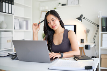 Obraz premium Asian woman sitting at home office desk and focusing on her laptop screen in a moment of contemplation.