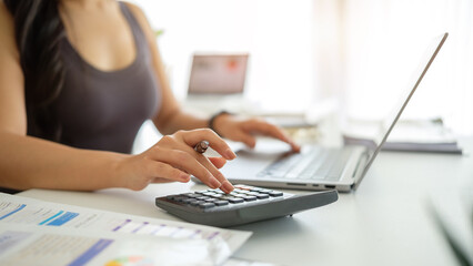 Close up of a woman calculating financial data using a calculator and laptop at a modern workspace.