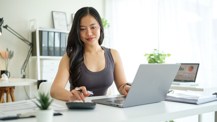 Confident asian woman using a laptop and calculator at home office desk, with charts, plants, and...