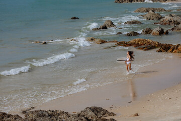 a young brunette woman in a sports swimsuit walks along the coastline with a surfboard in her hand