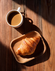 A top-down shot of a wooden table with a croissant, a cup of coffee, and soft morning light.