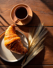 A top-down shot of a wooden table with a croissant, a cup of coffee, and soft morning light.