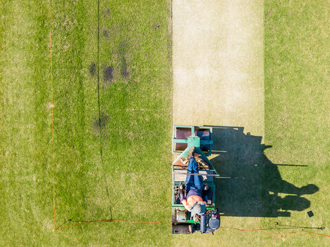 Looking down on a greens keeper riding a roller over a cricket pitch