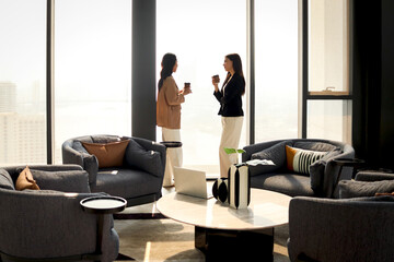 Two businesswomen have conversation during a break. Female business officers talking and drinking coffee together while standing in modern office. Businesspeople take a break at workplace.