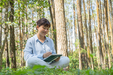 A young asian woman wearing a long-sleeve collared shirt and denim jeans pants is drawing using a digital tablet while sitting on the grass in a lush pine forest in the morning. Remote work concept