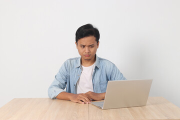 Young Asian man looking surprised while using a laptop. Shocked expression, sitting at a wooden desk isolated in white background. Concept of unexpected news and surprising online content