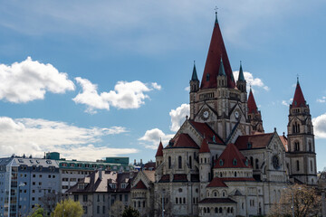 A wide-angle view of the majestic St. Francis of Assisi Church in Vienna, Austria, showcasing its distinctive red rooftops and Romanesque revival architecture against a partly cloudy blue sky.