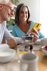 Vertical portrait of happy mature couple looking at mobile phone while enjoying healthy breakfast at home.